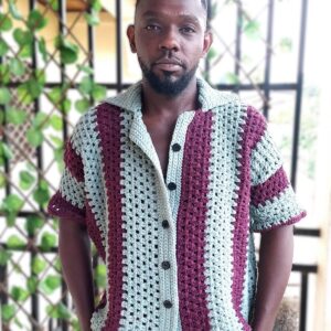 Homme portant une chemise boutonnée en crochet bicolore à rayures verticales vert sauge et prune, avec un col ouvert cubain et un pantalon blanc.