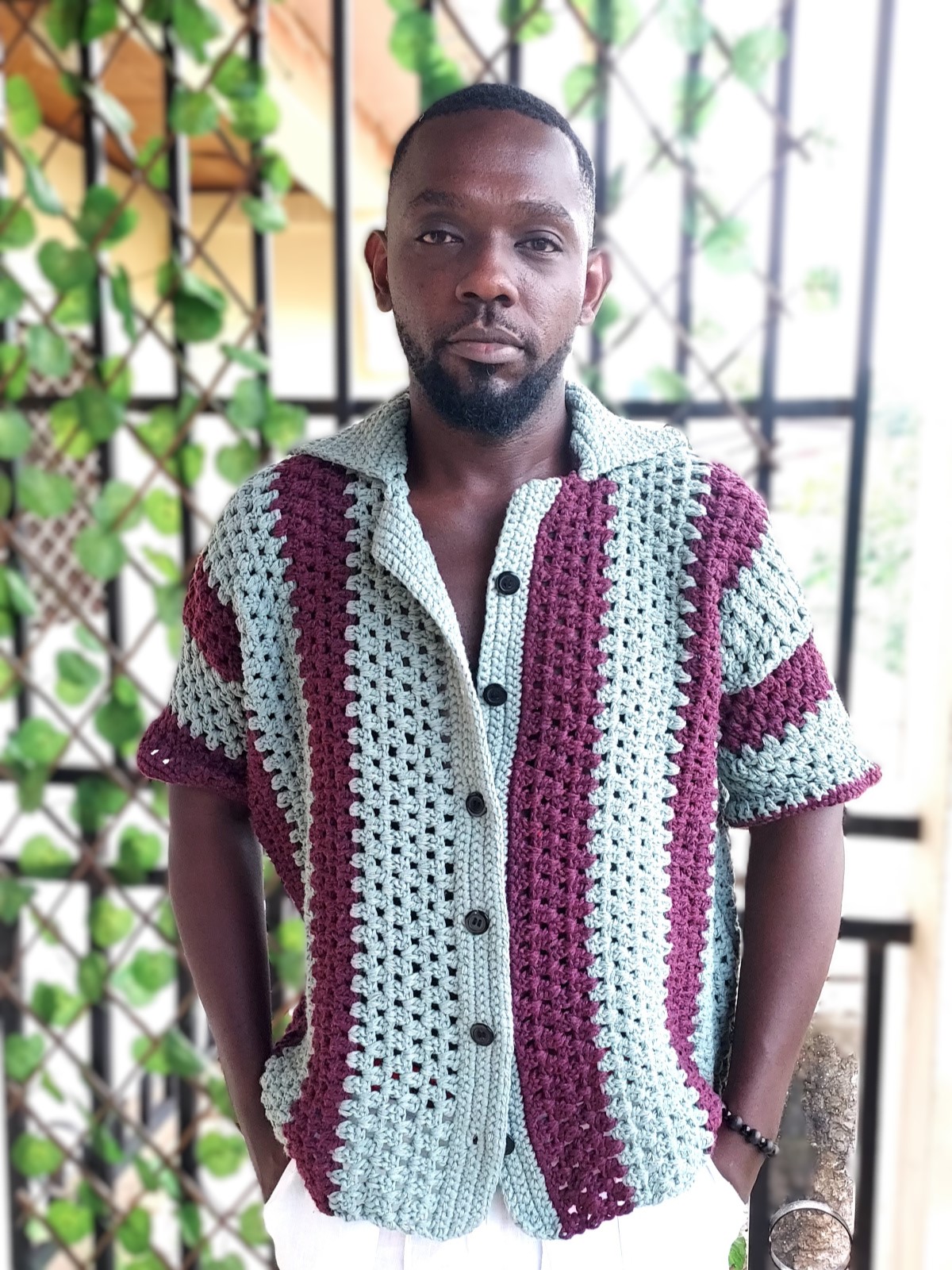 Homme portant une chemise boutonnée en crochet bicolore à rayures verticales vert sauge et prune, avec un col ouvert cubain et un pantalon blanc.